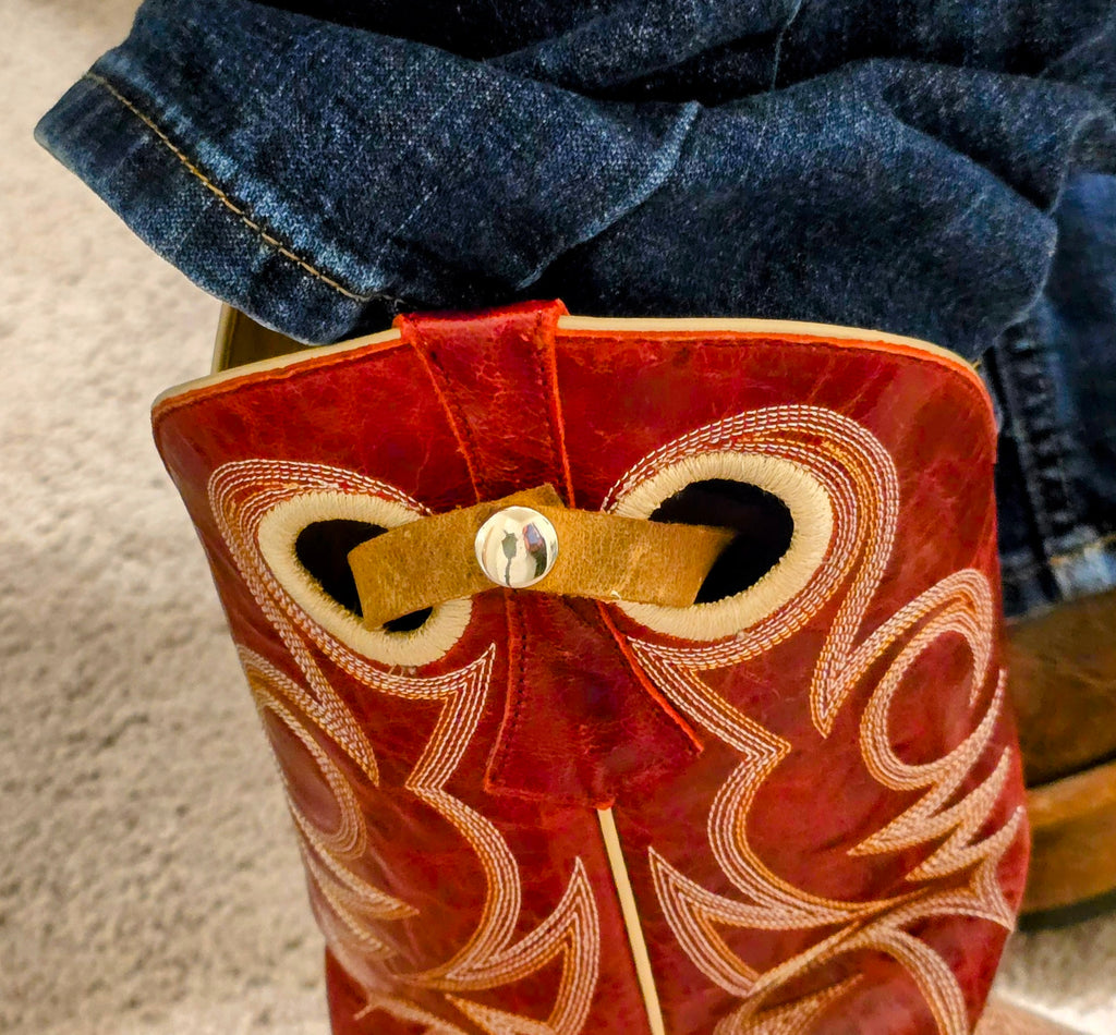 A close-up shot focusing on the red upper shaft of a cowboy boot, where the small brown leather strap of the sleeve is securely snapped around the internal boot pull. This view highlights how the sleeve attaches, keeping the card securely fastened to the boot pull while resting on the lower shaft, contrasting with the dark blue denim jeans.
