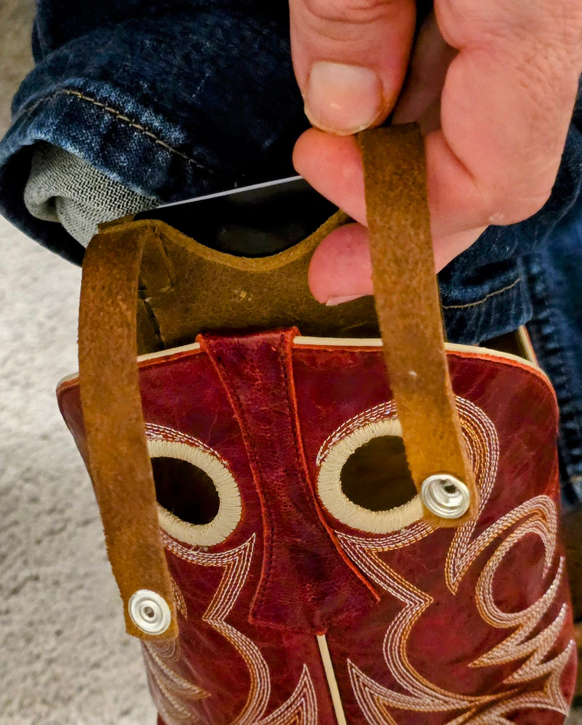 The human hand holds the open snap-handle of the rustic leather sleeve, which is cleverly looped through the pull hole on a highly stylized red cowboy boot. The card sleeve itself is positioned just below the pull, ready to be fastened, next to a rolled-up blue jean leg.
