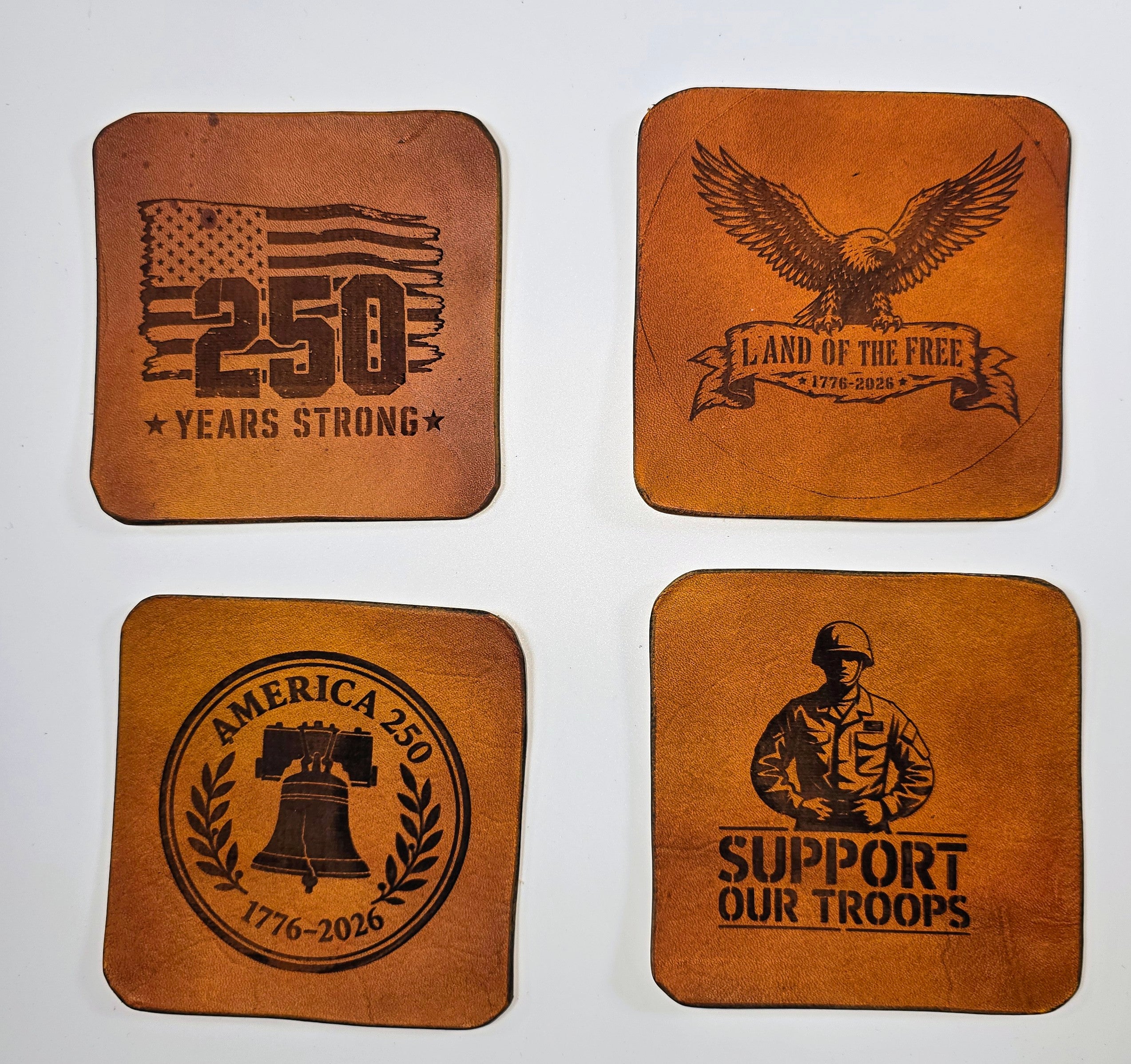 Flat lay of laser-engraved leather coasters from The Kilted Leatherworker, 4x4 inch square, rustic tan finish