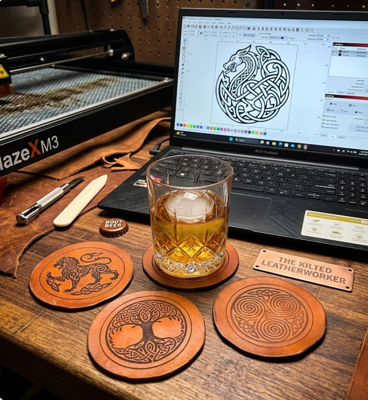 The Kilted Leatherworker Celtic leather coasters displayed on a workshop bench with a whiskey glass, laser engraver, and Celtic dragon design on laptop screen