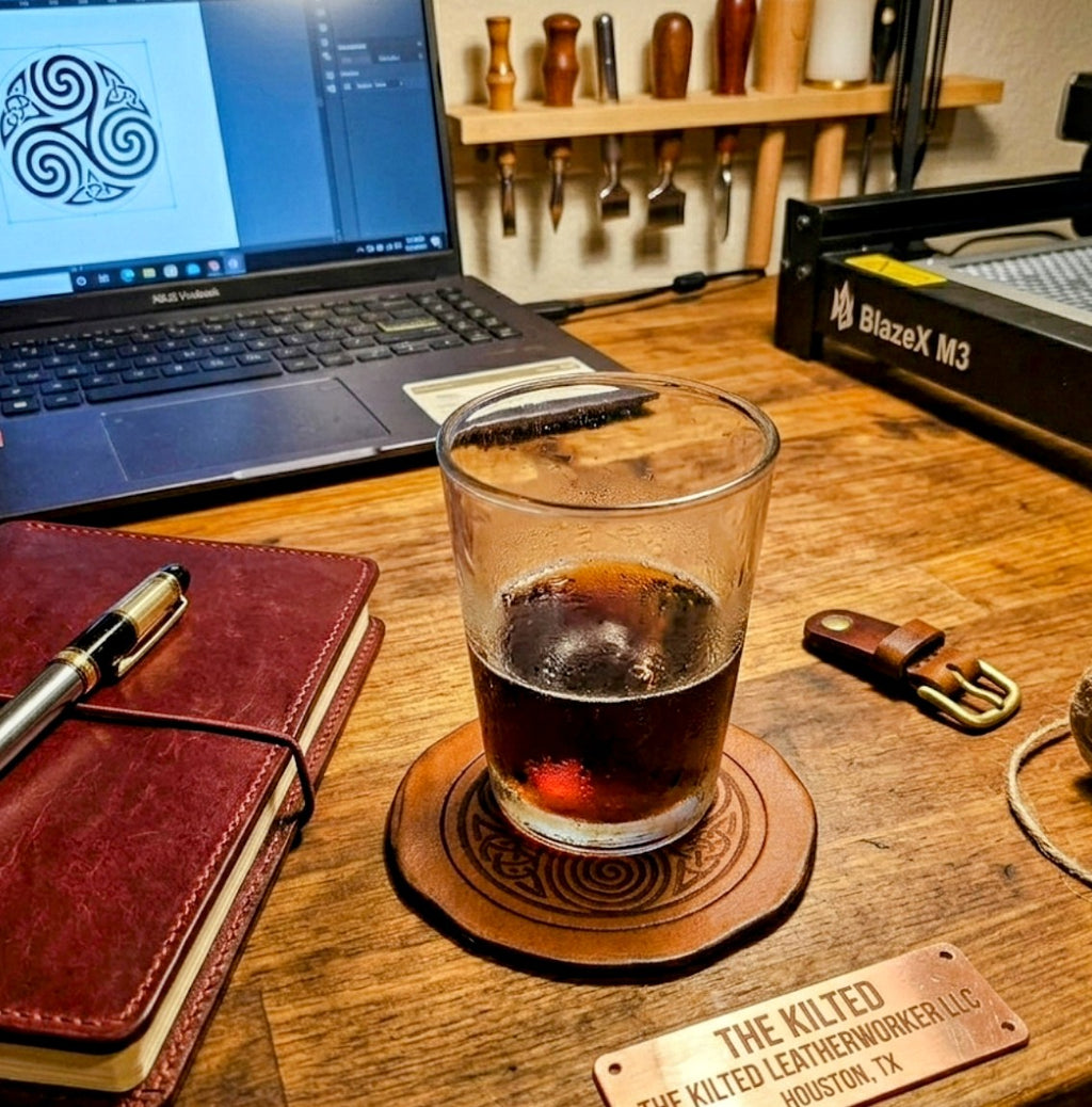 Handmade Celtic triskelion leather coaster on a craftsman's workbench with a whiskey glass, leather journal, and laser engraver in the background