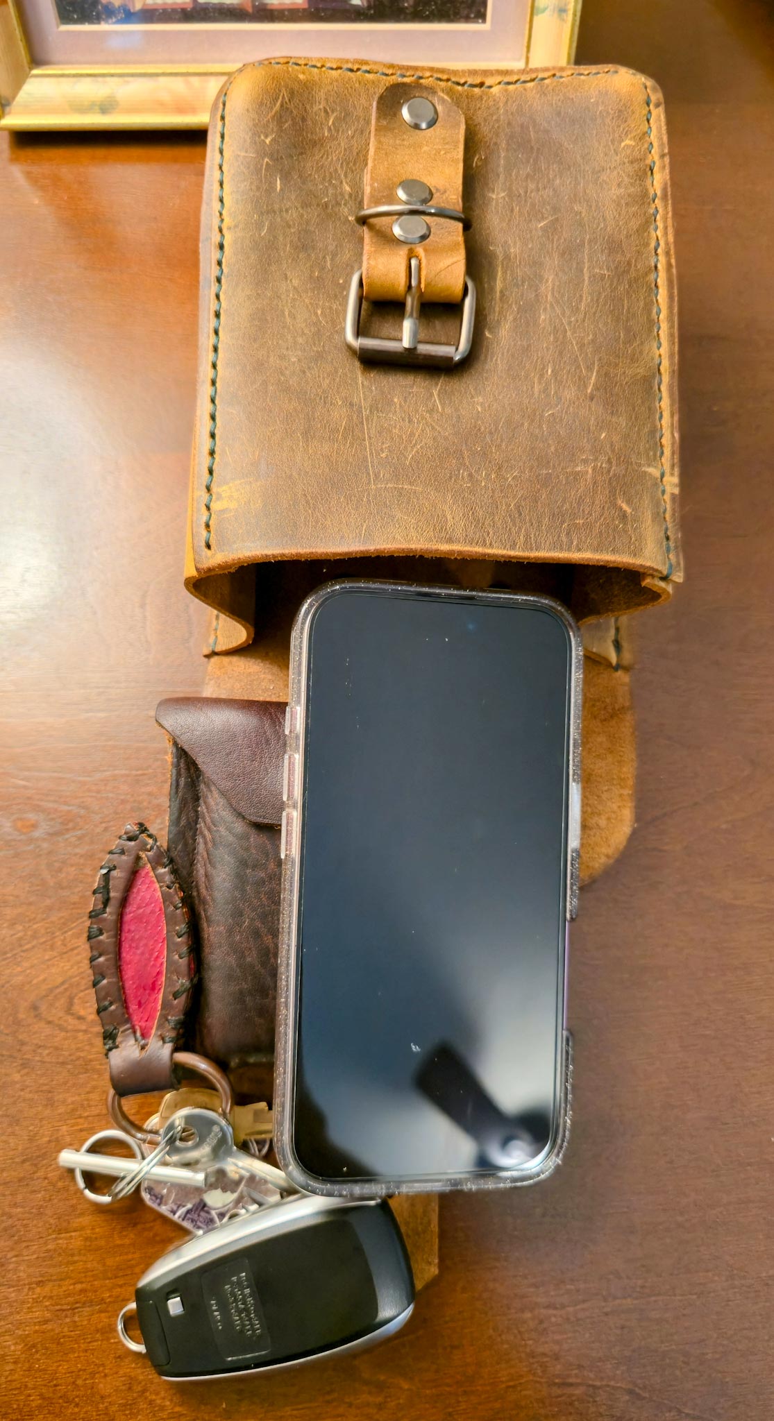 Leather belt pouch holding smartphone, wallet, and keys on wooden surface