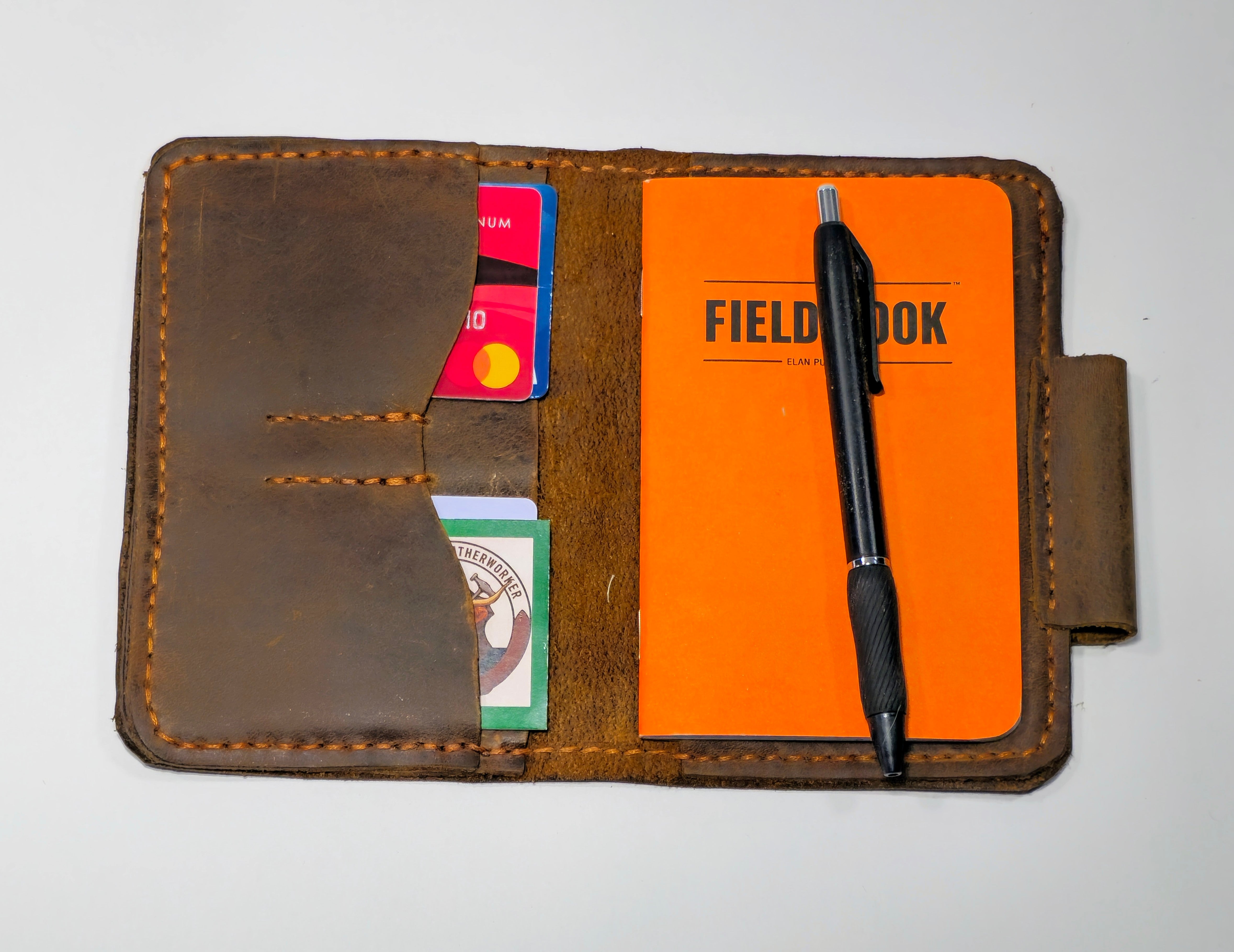 Brown leather wallet with orange field notebook, pen, and cards on a white background