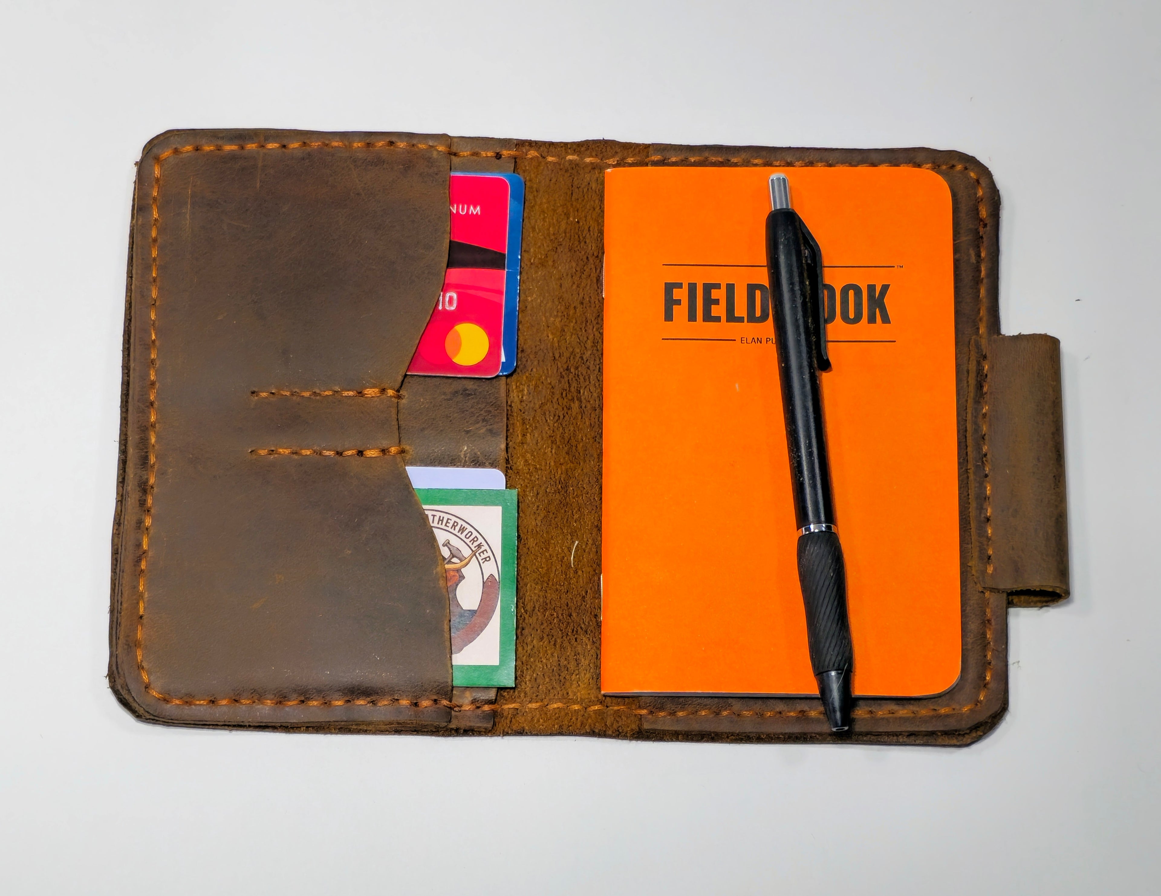 Brown leather wallet with orange field notebook, pen, and cards on a white background