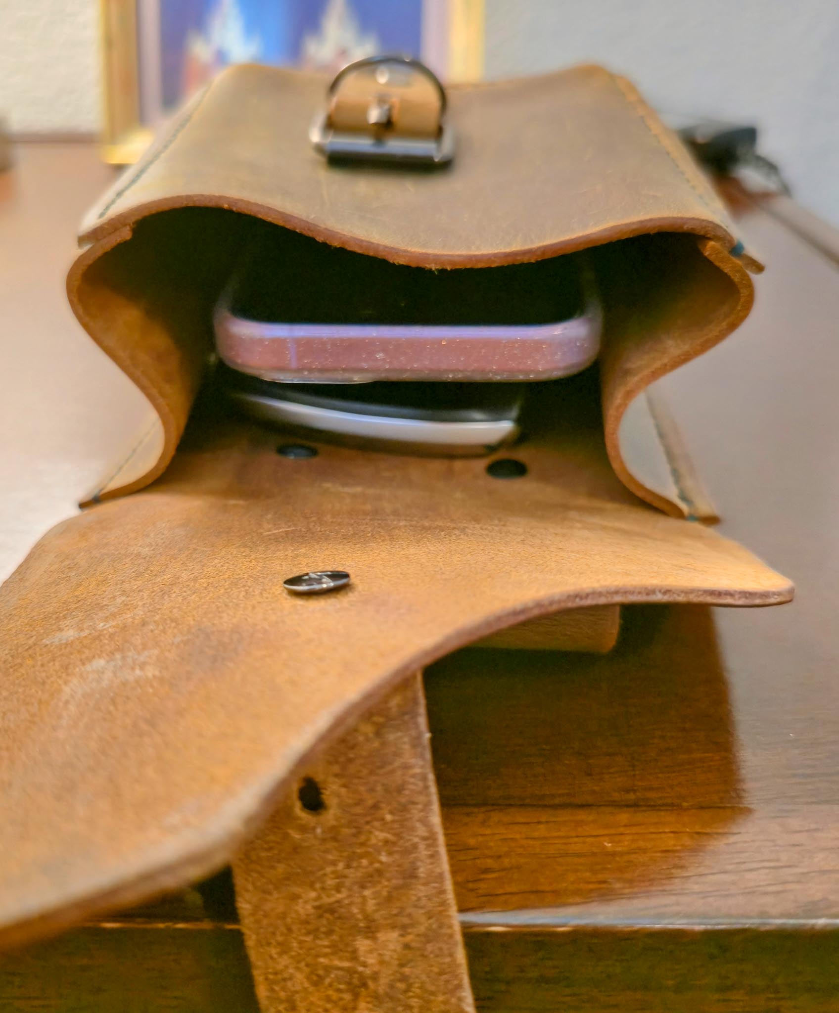 Brown leather belt pouch open with phone, keys, and wallet on wooden surface